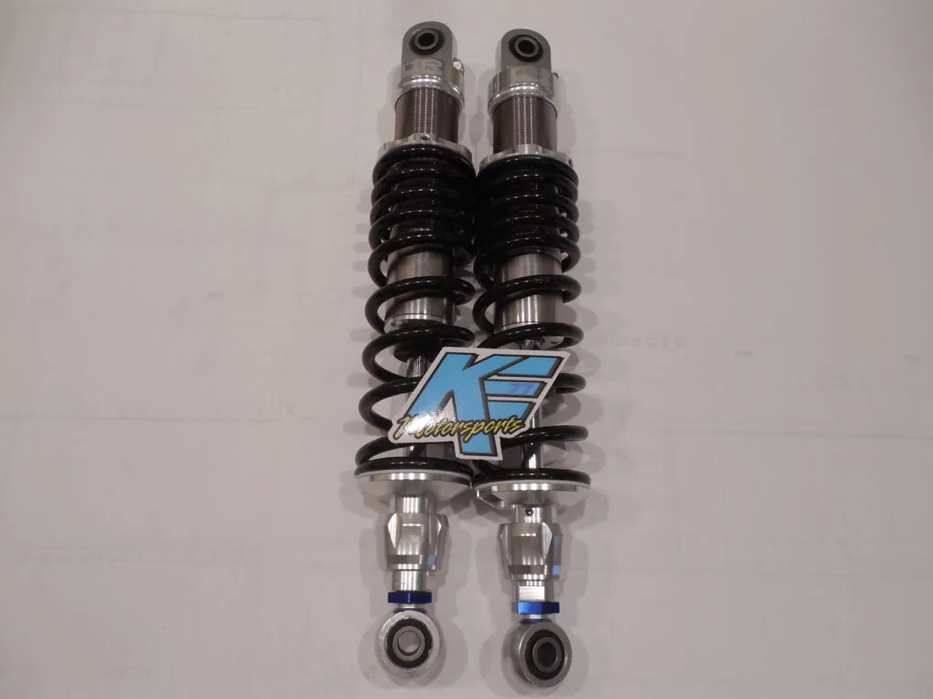 Pair of motorcycle shock absorbers with black coil springs, chrome bodies, and blue K logo badge mounted vertically on white background