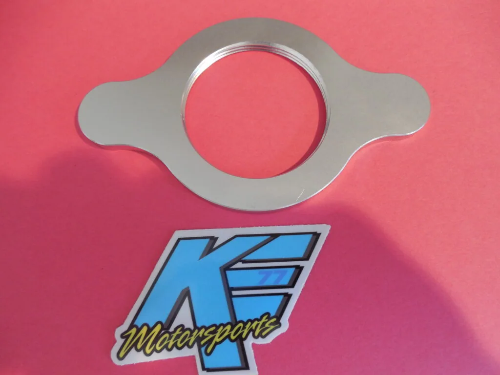 Gray metal circular gasket with two ear-like extensions on a pink background above KE Motorsports logo