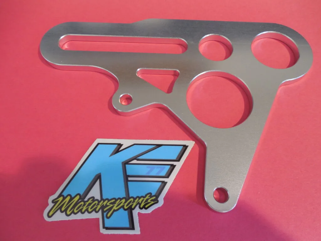 Gray metal mounting bracket with circular and slot-shaped cutouts on pink background with K.E. Motorsports logo sticker.