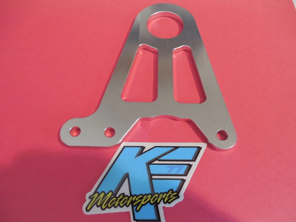 Gray metal bracket with circular opening and two rectangular slots mounted on pink surface with blue motorsports logo badge