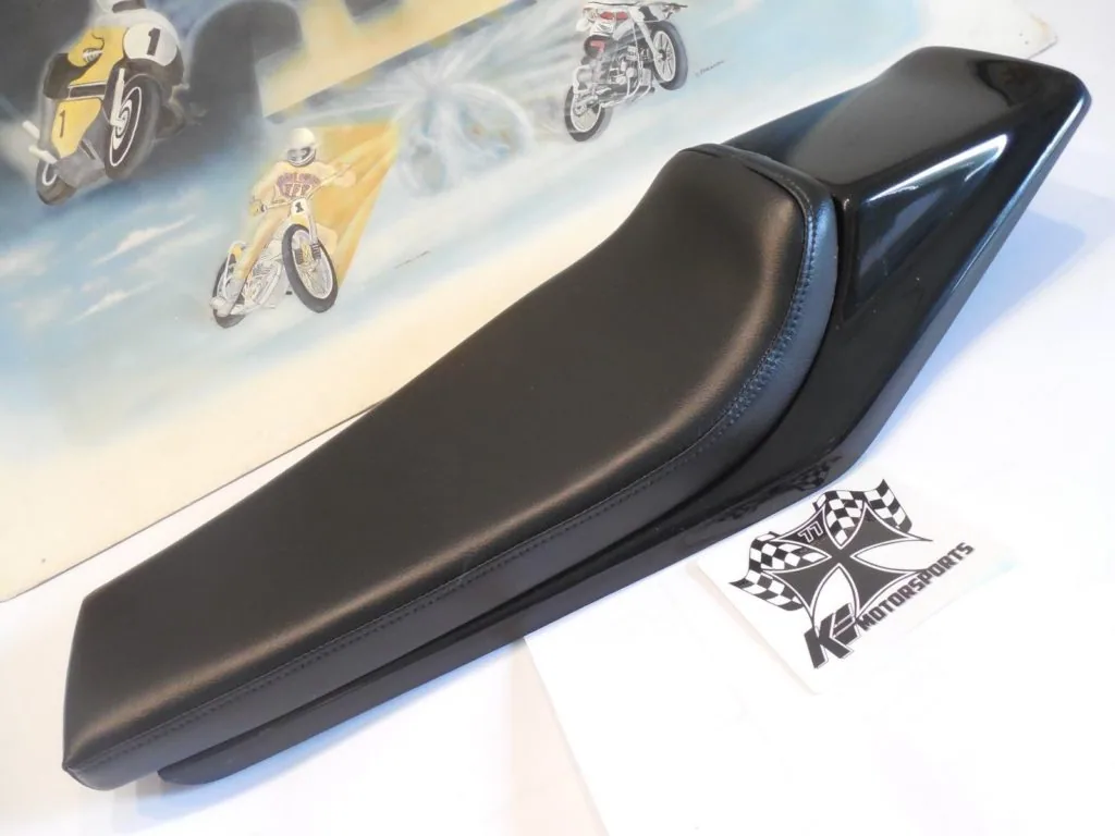 Black motorcycle seat with blue stitching displayed on white surface next to K.E. Motorsports logo and motorcycle artwork backdrop