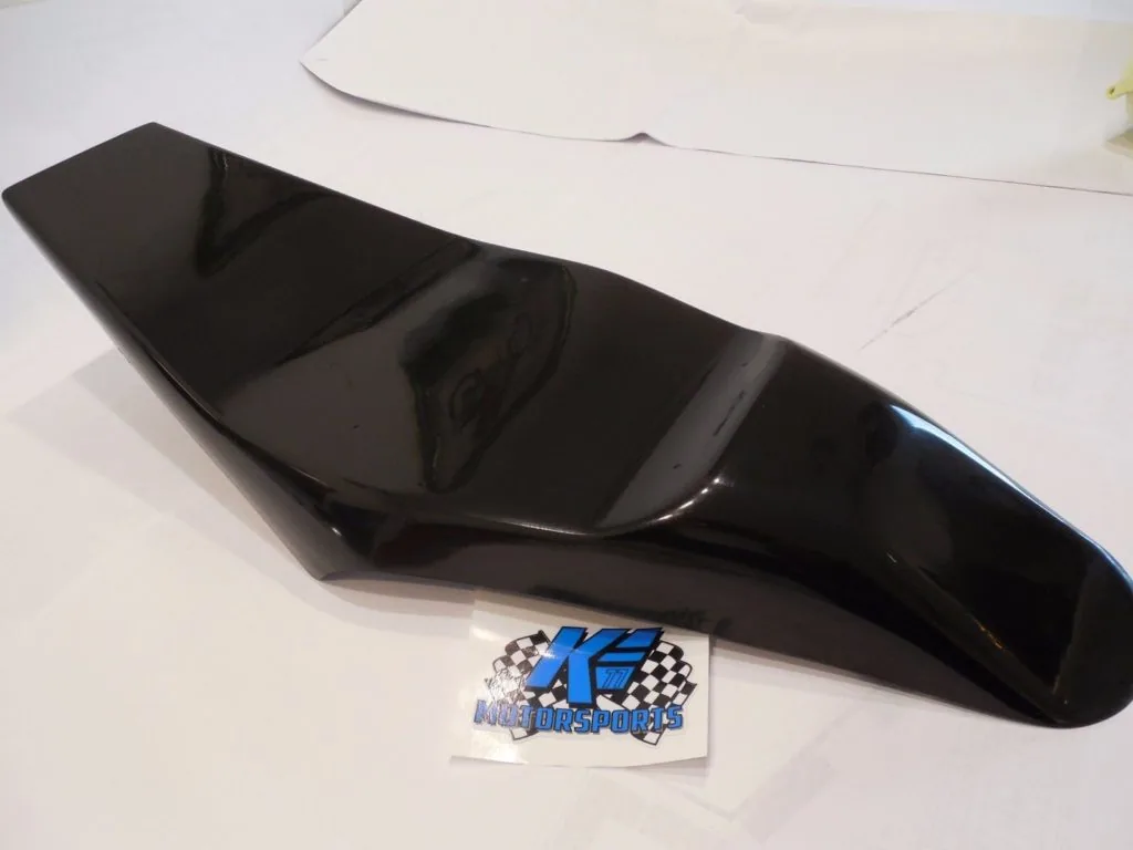 Black glossy motorcycle seat with dual panels on white surface next to K.E. Motorsports sticker
