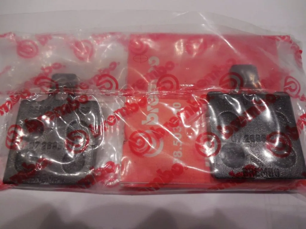 Two dark metal brake pads in clear plastic packaging with red patterned backing