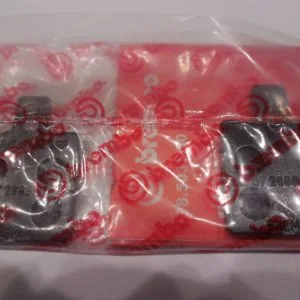 Two dark metal brake pads in clear plastic packaging with red patterned backing