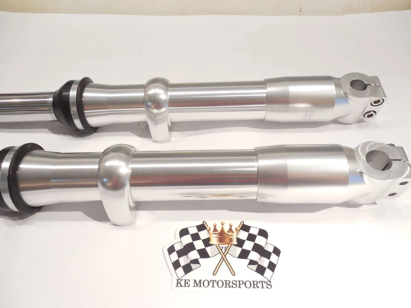 Two polished silver suspension damper shocks with white adjustment knobs and black seals on white background with KE Motorsports logo.