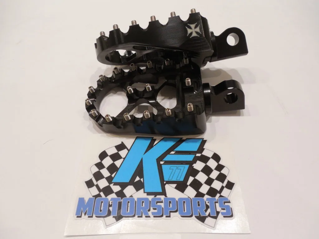 Black motorcycle footpegs with serrated surfaces and mounting brackets displayed above K.E. Motorsports logo with checkered flags