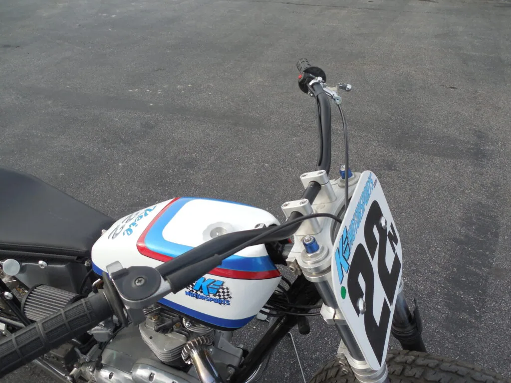 Overhead view of motorcycle with white tank featuring red and blue stripes and number plate mounted on handlebars