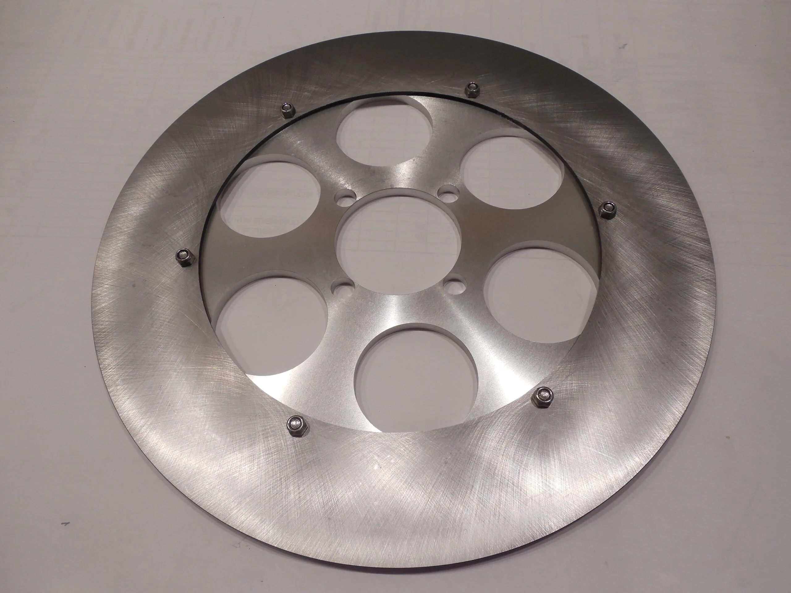 Brushed metal circular component with six circular cutouts arranged around a central opening, secured with small bolts.