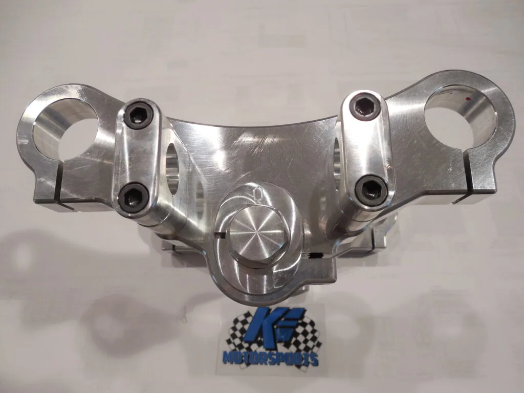 Polished aluminum motorcycle handlebar clamp with dual mounting points and circular emblem detail, shown with K.E. Motorsports logo