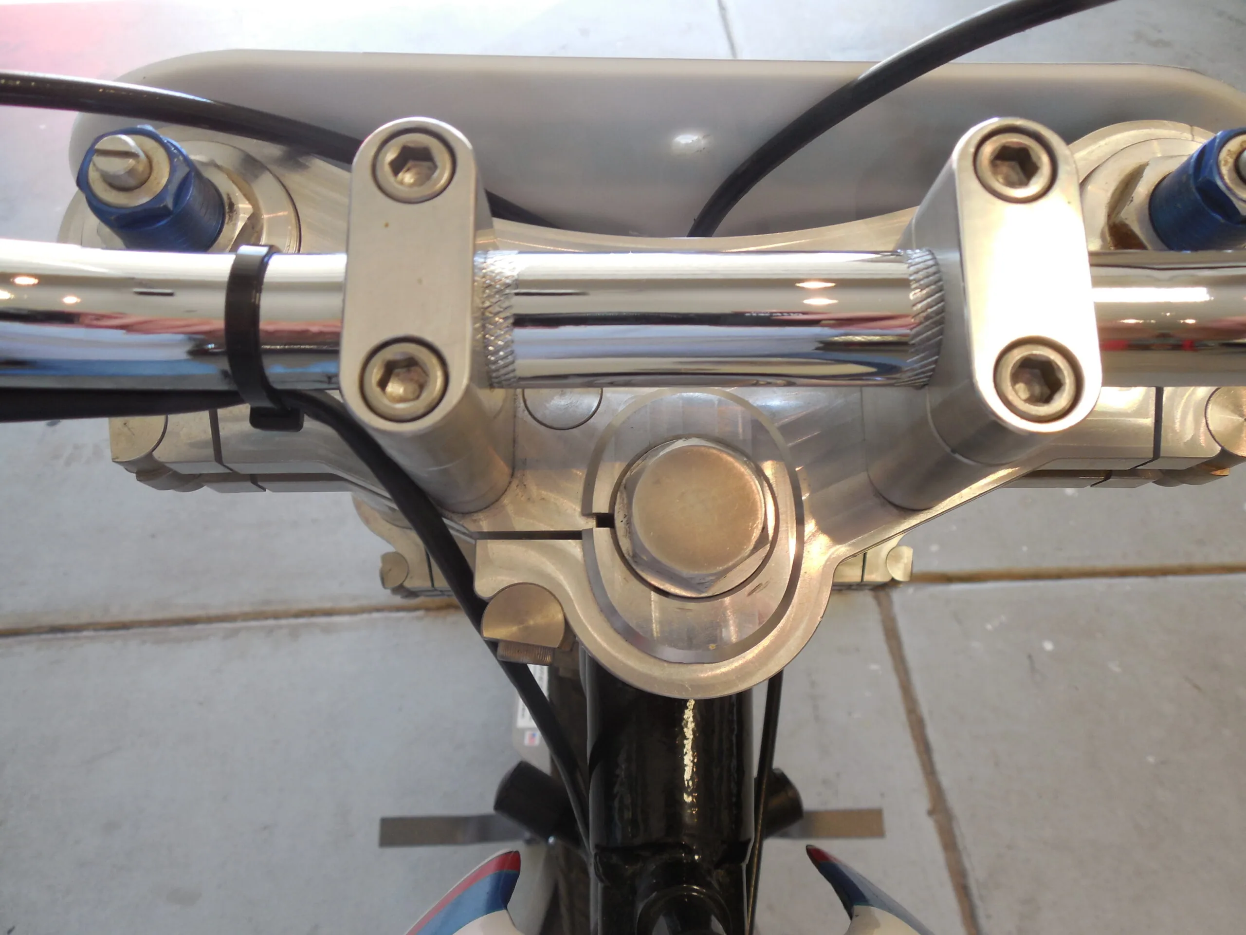 Motorcycle handlebar with chrome risers, white mounting brackets, and black cables against gray fairing background.
