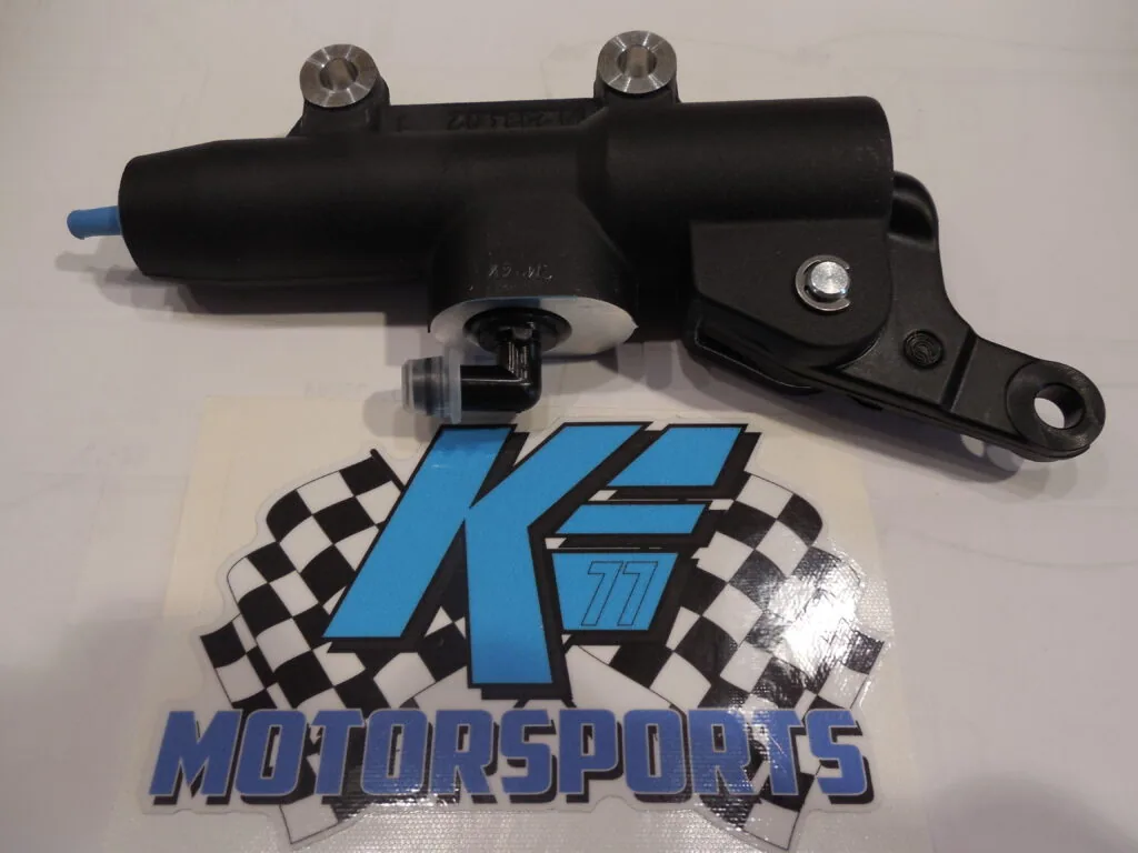 Black hydraulic brake master cylinder with dual pistons and blue inlet line positioned above K.E. Motorsports logo with checkered flags