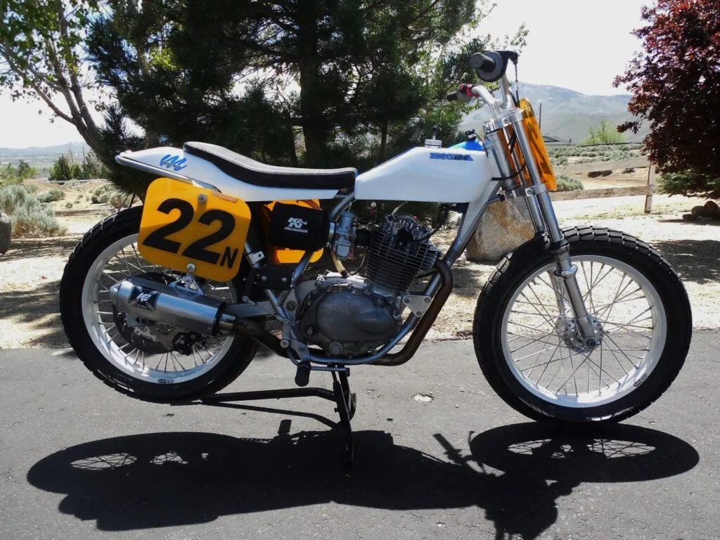 Flat track motorcycle with a yellow number 22 plate, white tank and black seat, standing in the sun